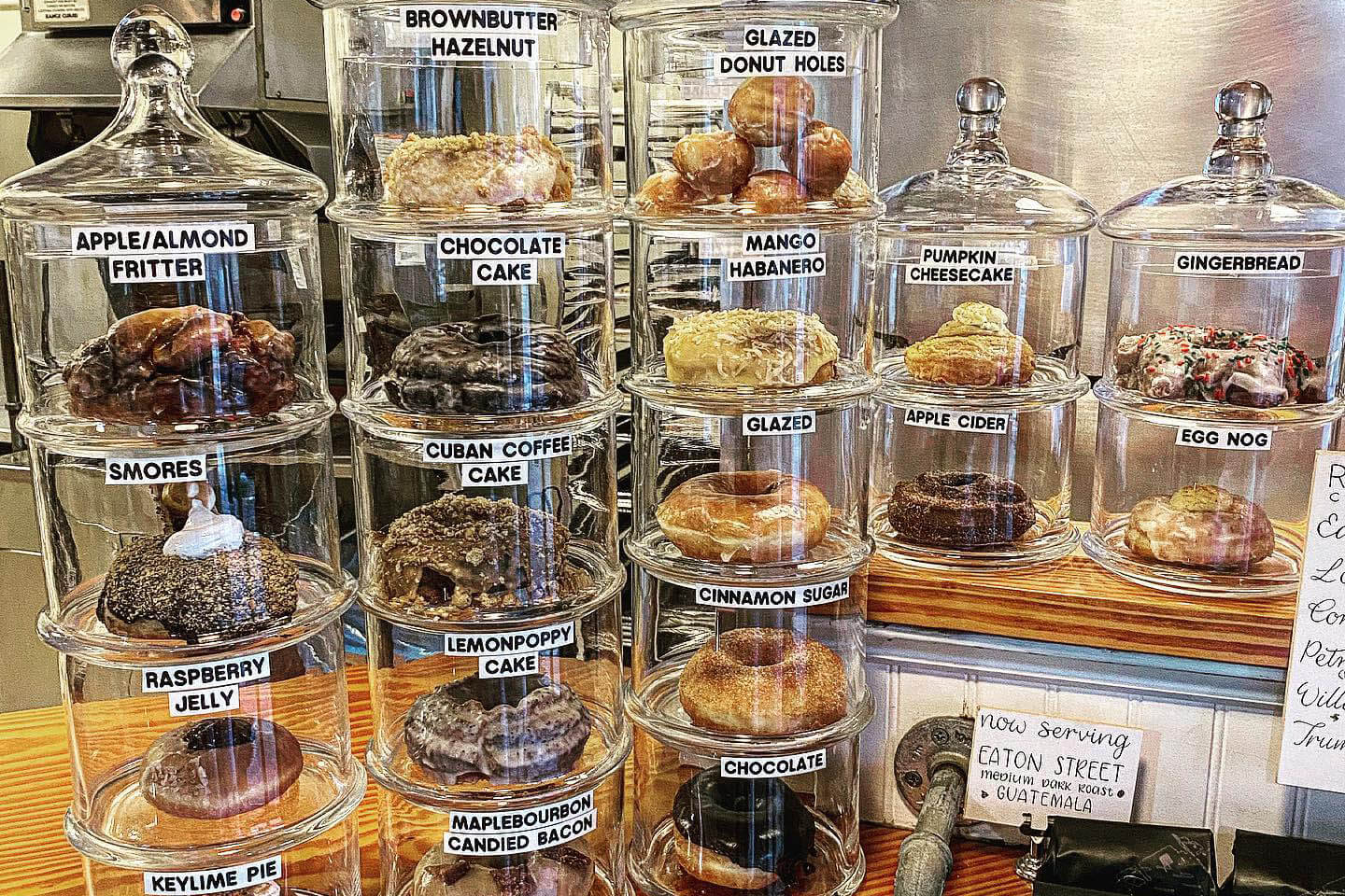 Donuts in glass containers. Labels on containers read Apple/Almond Fritter, Smores, Raspberry Jelly, Kyelime Pie, Bownbutter Hazelnut, Chocolate Cake, Cuban Coffee Cake, Lemon Poppy Cake, Maple Bourbon Candied Bacon, Glazed Donut Holes, Mango Habanero, Glazed, Cinnamon Sugar, Chocolate, Pumpkin cheesescake, Apple Cider, Gingerbread, and Egg Nog. 