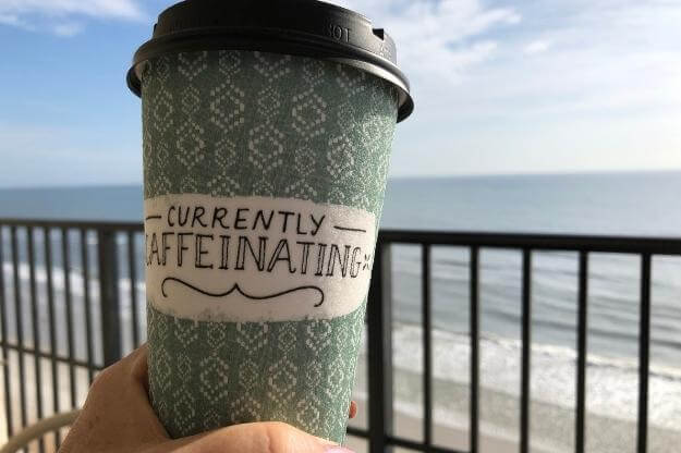 Coffee cup by the beach. Lettering on the cup says Currently Caffeinating. 