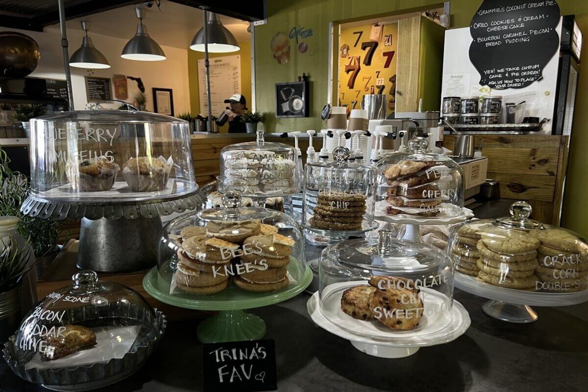 Baked goods on display with wording on the tops of each container. Visible wording includes Chocolate Chip Sweet, Cracked Corn Double, Bee's Knee's, Blueberry muffins, bacon checddar chive, and chocolate chip. 